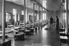 An Army barracks room with rows of cots. A soldier wearing a pre-World-War-Two uniform stands at parade rest while another soldier polishes the floor with an electric buffer.